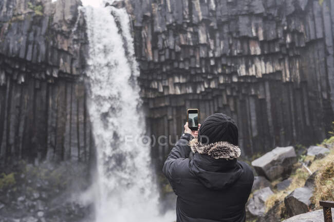Rückansicht eines unkenntlich gemachten männlichen Reisenden in warmer Oberbekleidung und Hut mit Smartphone beim Fotografieren des malerischen Wasserfalls Svartifoss, der während einer Reise in Island durch Basaltklippen fließt — Stockfoto
