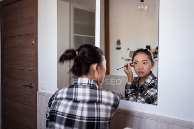Focused charming ethnic female applying eyebrow pencil while doing makeup and looking in mirror — Stock Photo