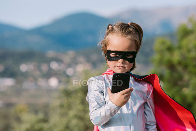 Chica segura de sí mismo en disfraz de superhéroe máscara con la capa de navegación en el teléfono celular - foto de stock