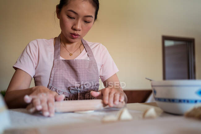 Снизу женщина в фартуке катит тесто на столе, пока готовит пельмени с мясом на кухне — стоковое фото