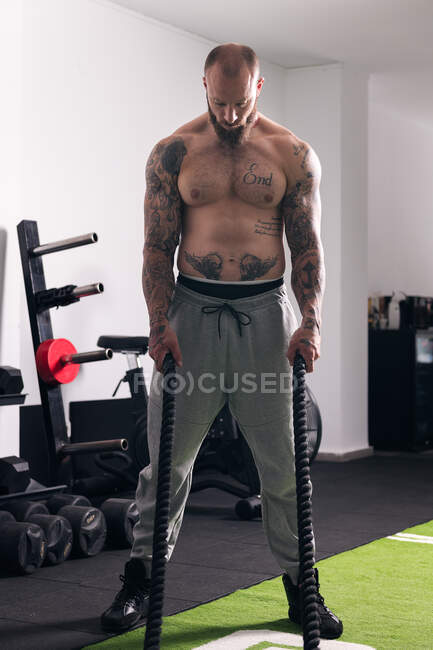 Concentrated shirtless sportsman standing with ropes during functional workout in gym and looking down in daytime — Stock Photo
