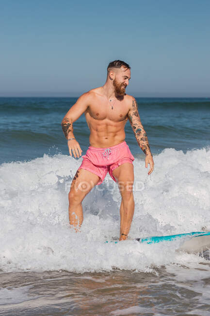Homem ativo em shorts de natação em pé na prancha de surf enquanto navega em mar ondulado em resort tropical no dia ensolarado de verão — Fotografia de Stock
