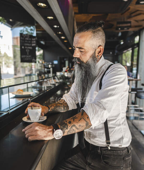 Side view of serious bearded businessman with tattoo in classy outfit drinking coffee while sitting at counter in modern cafeteria — Stock Photo