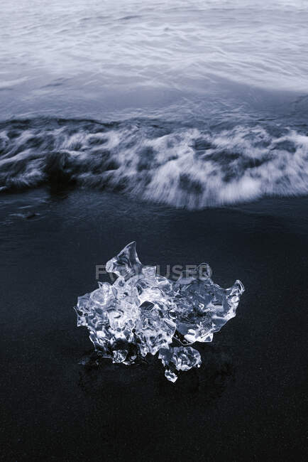 Сверху кусок блестящего льда на черном песчаном пляже, омываемом волнистым океаном в облачный день в Исландии — стоковое фото