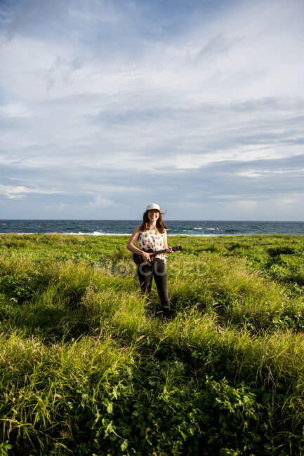 Щаслива жінка-музикантка з коричневим волоссям в повсякденному одязі, що стоїть на укулеле, дивлячись на камеру в полі з зеленою травою проти пагорбів на сонячному світлі — стокове фото