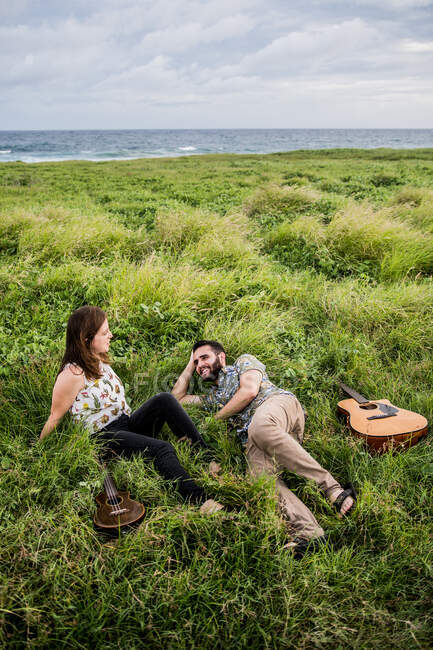 Amici felici musicisti con chitarra e ukulele seduti su erba verde sulla costa vicino all'oceano in natura durante il giorno — Foto stock