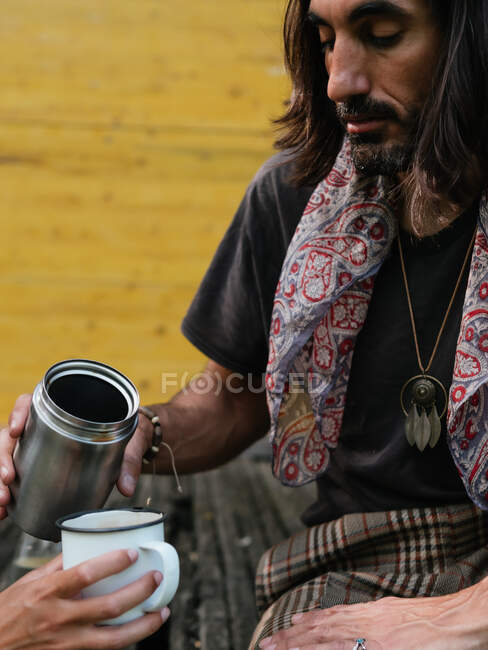 Hippie-Mann gießt Kaffee oder Tee aus einer Thermoskanne in eine Tasse, die eine anonyme Frau auf gelbem Holzgrund hält — Stockfoto