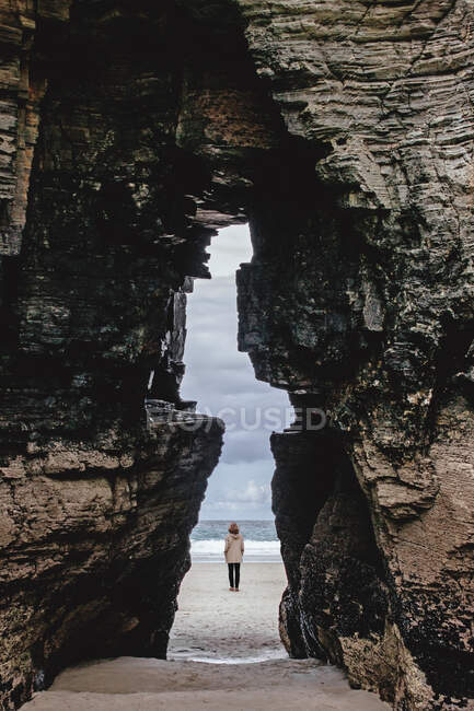 View of distant anonymous traveler in outerwear through massive stony formation on sandy coast near sea in nature of Spain — Stock Photo