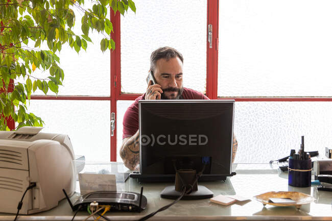 Hombre adulto serio sentado en la mesa mientras habla por teléfono y navega en el ordenador en el espacio de trabajo de luz cerca de ventanas y plantas verdes - foto de stock