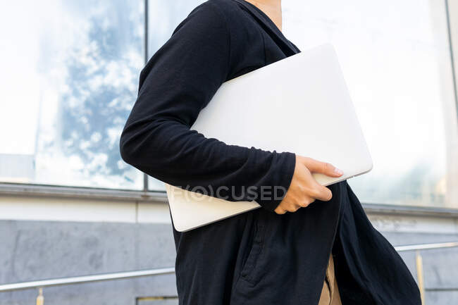 Вид збоку анонімного студента-чоловіка в повсякденному одязі з сучасним нетбуком в руці, що йде по вулиці біля сучасної будівлі — стокове фото