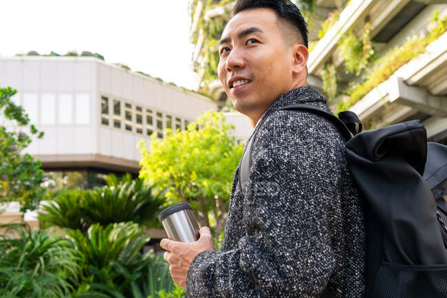 Side view of dreamy young Asian businessman with rucksack and tumbler looking away against plants and urban houses — Stock Photo