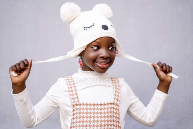 Cheerful African American little girl in stylish clothes and funny hat standing with raised arms and looking away against gray background — Stock Photo