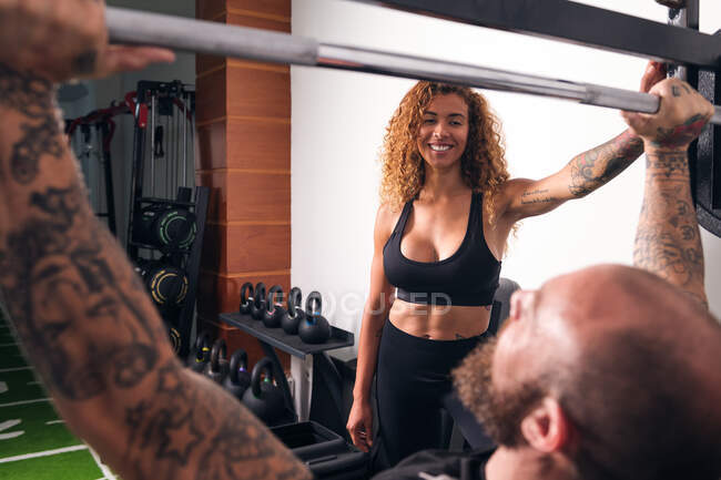 Вид сзади спортсмен с татуировками, поднимающий штангу с тяжелыми грузами во время функциональной тренировки с инструктором женского пола в тренажерном зале — стоковое фото