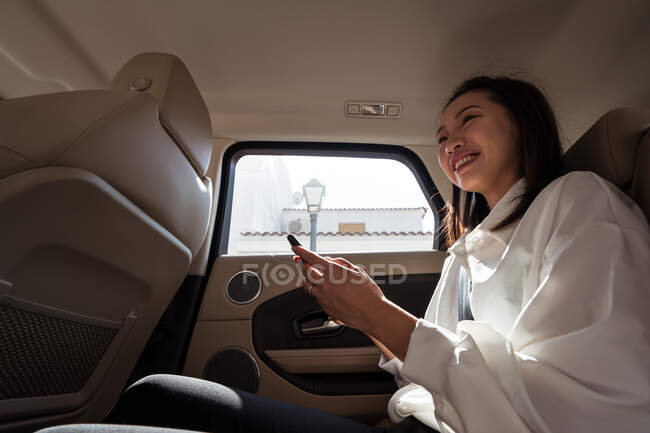 Vista lateral de pasajera asiática en ropa formal navegando por teléfono celular mientras está sentada en el asiento trasero en taxi y mirando hacia otro lado con interés - foto de stock