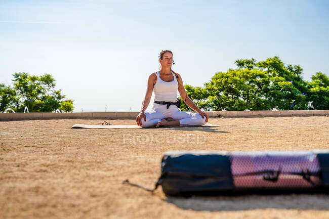 Tranquilo professor de ioga feminino com microfone sentado no homem e realizando Sukhasana contra árvores verdes e céu azul sem nuvens no dia ensolarado — Fotografia de Stock