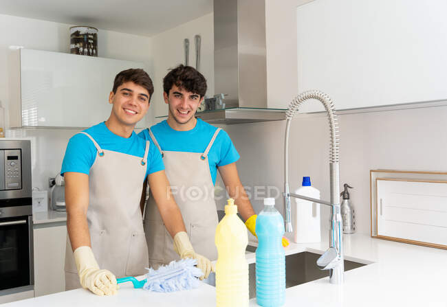 Позитивні чоловічі прибиральники у формі та гумові рукавички посміхаються та дивляться на камеру, коли працюють на сучасній кухні вдень. — стокове фото