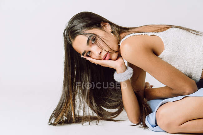 Vista lateral de la joven hispana con el pelo largo tocando la cara mientras se inclina hacia adelante y mirando a la cámara en el suelo - foto de stock