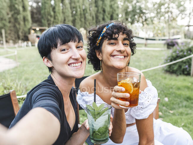 Entzückte multirassische beste Freundinnen mit Cocktailgläsern, die im Sommerpark chillen und ein Selfie mit dem Smartphone machen — Stockfoto