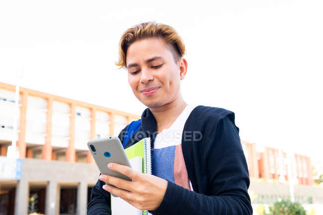 Estudante masculino alegre no desgaste casual com copybooks mensagens de texto no celular moderno, enquanto está perto do edifício da universidade durante os estudos — Fotografia de Stock