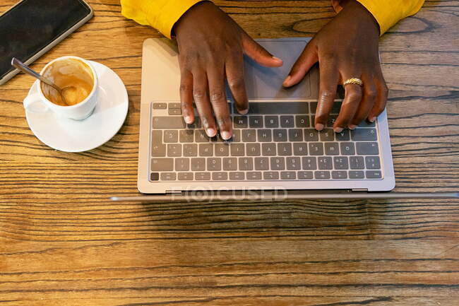 From above of crop anonymous African American person browsing modern netbook while sitting at wooden table with empty cup of coffee in cafeteria — Stock Photo