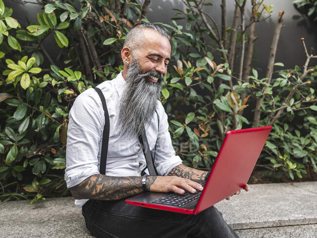 Freelancer masculino hipster positivo em uso formal navegando netbook moderno enquanto sentado na fronteira de pedra perto de plantas verdes na rua durante o trabalho on-line — Fotografia de Stock