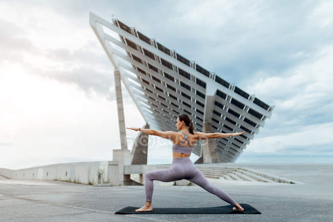 Cuerpo completo de mujer deportiva en ropa deportiva practicando la postura del guerrero mientras entrenaba en la calle cerca del panel solar contra el cielo nublado - foto de stock