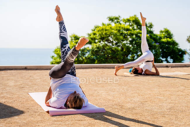 Flexible Frau in Aktivkleidung führt Eka Pada Sarvangasana auf Matte auf trockenem Boden während einer Yoga-Stunde im Park vor grünen Bäumen und wolkenlosem blauen Himmel im Sonnenlicht vor — Stockfoto