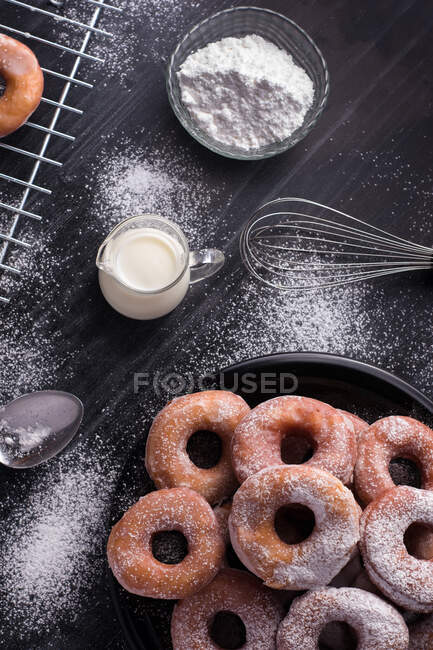 Beignets frits sucrés servis sur une assiette près d'une grille de refroidissement en métal et une cruche de lait sur une table noire avec du sucre en poudre — Photo de stock