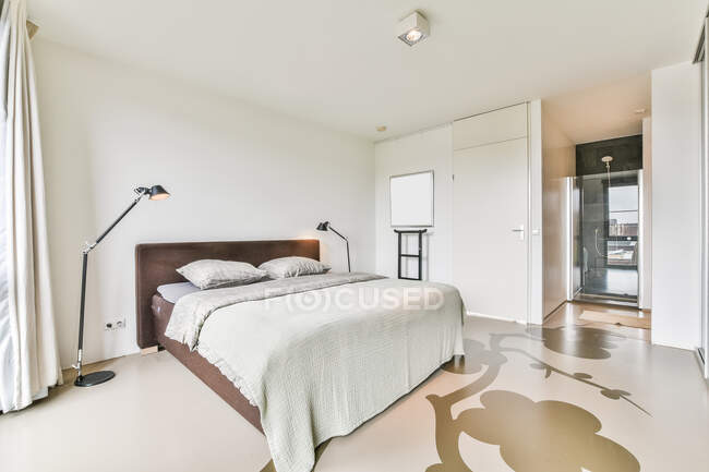 Уютная кровать с подушками и одеялом, размещенная в стильной спальне с занавесом на окнах и светящейся черной лампой — стоковое фото
