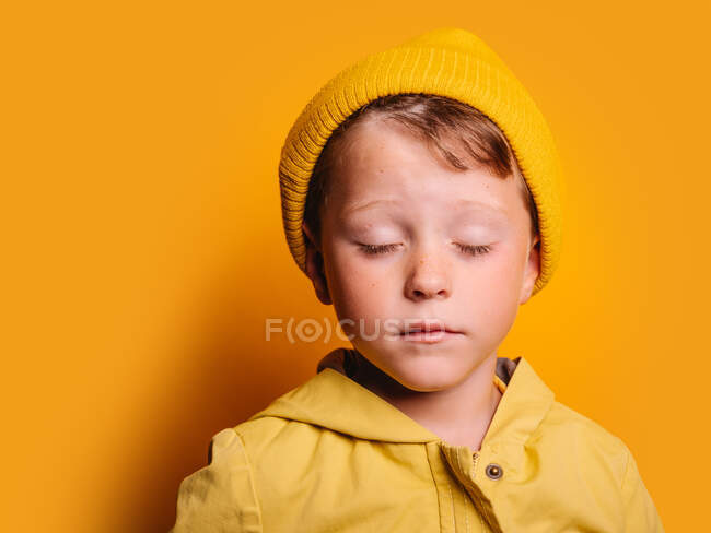 Niño poco emocional en impermeable de moda y gorro sombrero de pie con los ojos cerrados contra el fondo amarillo en el estudio - foto de stock