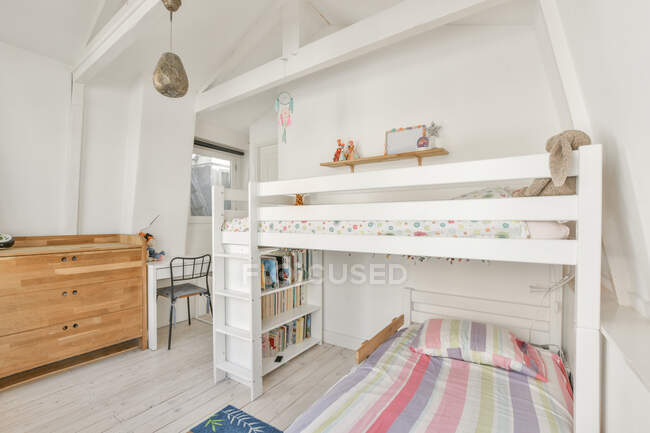 Біле двоярусне ліжко з книжковими полицями та барвистим килимом біля дверей у світлій сучасній дитячій спальні — стокове фото