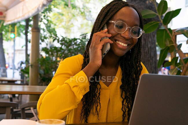 Alegre freelancer afroamericana teniendo conversación telefónica mientras está sentada en la mesa con netbook y notebook en la cafetería al aire libre durante el trabajo remoto - foto de stock