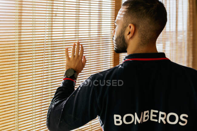 Вид сбоку с урожая на молодого бородатого этнического мужчину в спортивной куртке пожарного, смотрящего в окно и трогающего жалюзи пальцами при ярком солнечном свете — стоковое фото