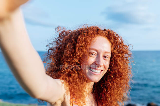 Feliz mujer de pelo rizado brazos extendidos mirando a la cámara mientras toma autorretrato en el teléfono inteligente en la costa de la colina de la orilla del mar - foto de stock