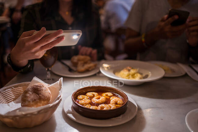 From above of crop anonymous woman taking photo of delicious food with saucy shrimps on smartphone while having dinner with boyfriend in restaurant — Stock Photo