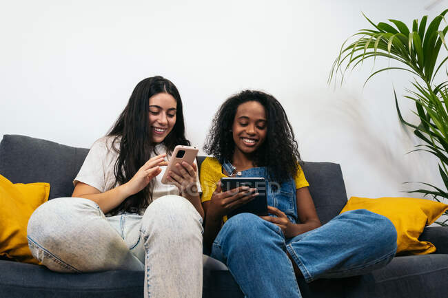 Jóvenes diversas amigas en ropa casual sonriendo mientras se sientan en el sofá navegando en el teléfono inteligente en la sala de estar en casa - foto de stock