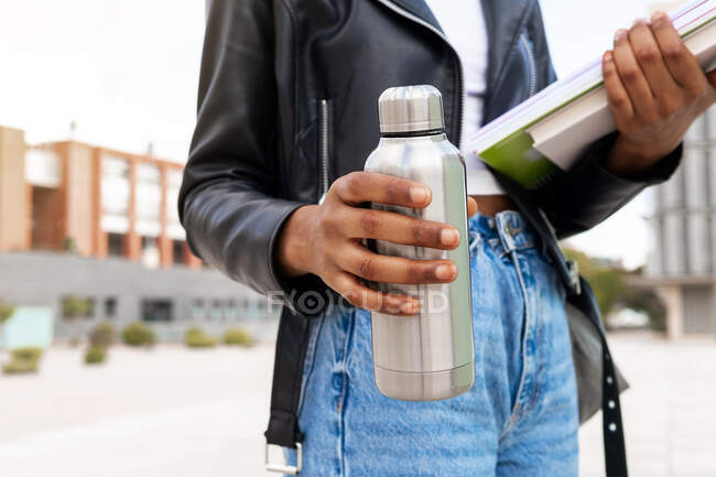 Estudiante afroamericana anónima con termos y un montón de libros de texto en la calle cerca del edificio de la universidad - foto de stock