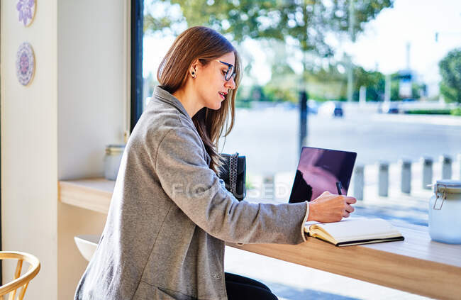 Вид сбоку сосредоточенной женщины-предпринимателя в очках, делающей заметки в планировщике во время работы на ноутбуке в кафе — стоковое фото