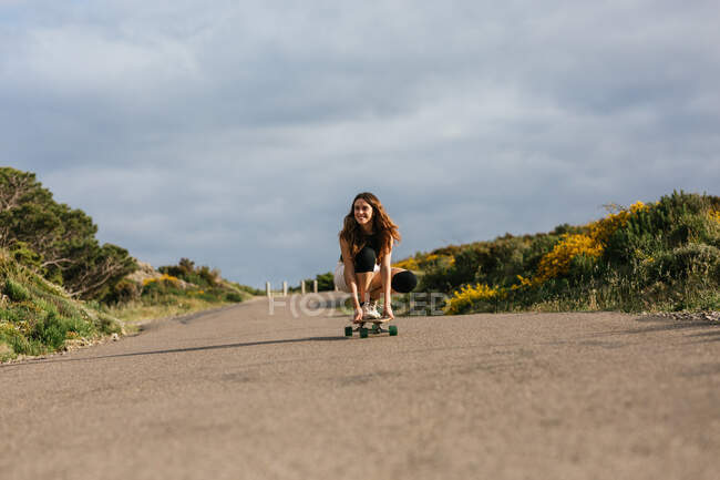 Cuerpo completo de feliz joven hembra en rodilleras montando longboard en camino de asfalto entre colinas bajo el cielo nublado - foto de stock