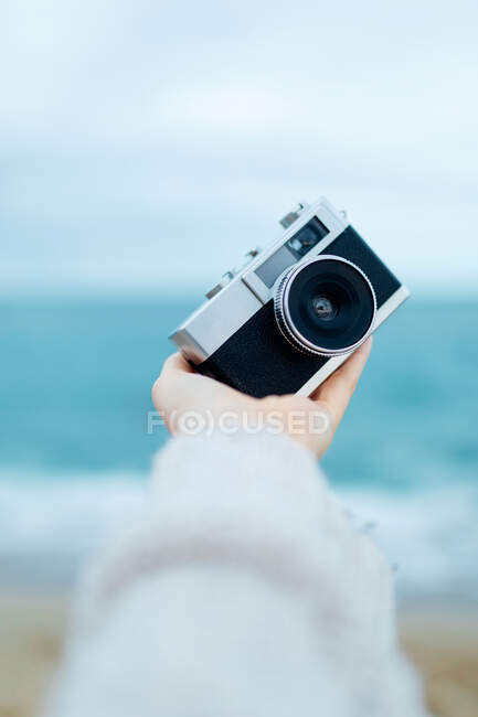 Crop unrecognizable female traveler demonstrating retro photo camera while standing on beach washed by sea waves — Stock Photo