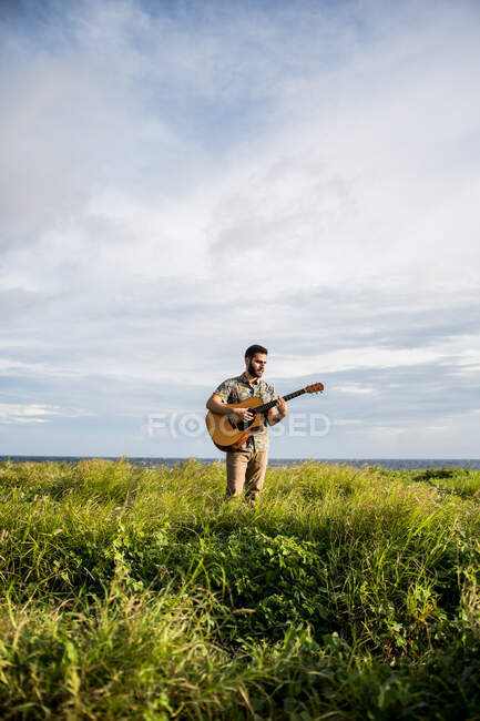 Calm man musician in casual clothes standing among green grass on coast of ocean and playing acoustic guitar in summertime in daylight — Stock Photo