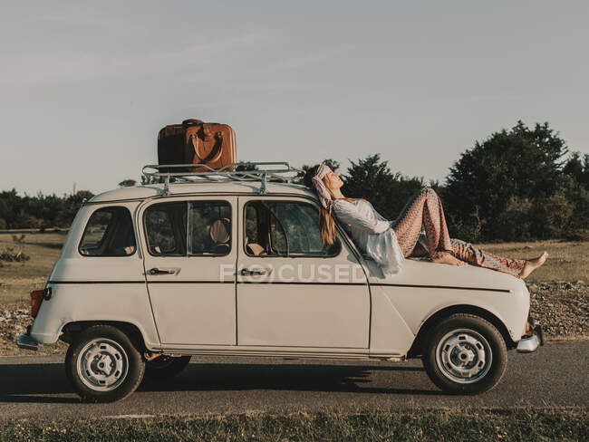 Ganzkörper-Seitenansicht einer barfüßigen Hippie-Frau im Boho-Outfit auf einem weißen Retro-Auto mit Koffer während einer Reise in der Natur — Stockfoto