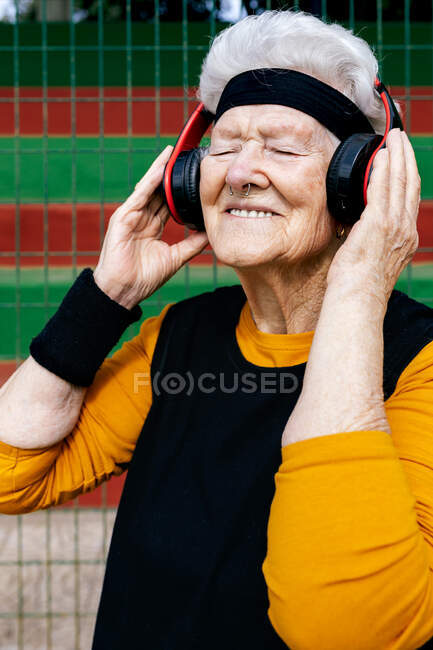 Contenido hembra madura con la nariz perforada en desgaste activo escuchando canciones en auriculares mientras está de pie campo de deportes cerca de la red - foto de stock