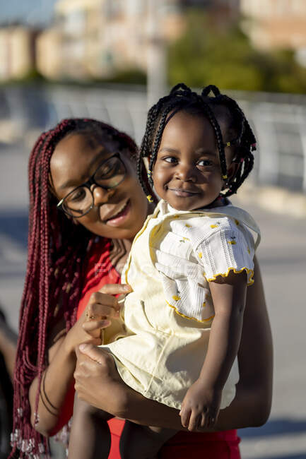 Mère afro-américaine joyeuse avec des tresses rouges debout avec une petite fille positive sur les mains dans la rue au soleil — Photo de stock