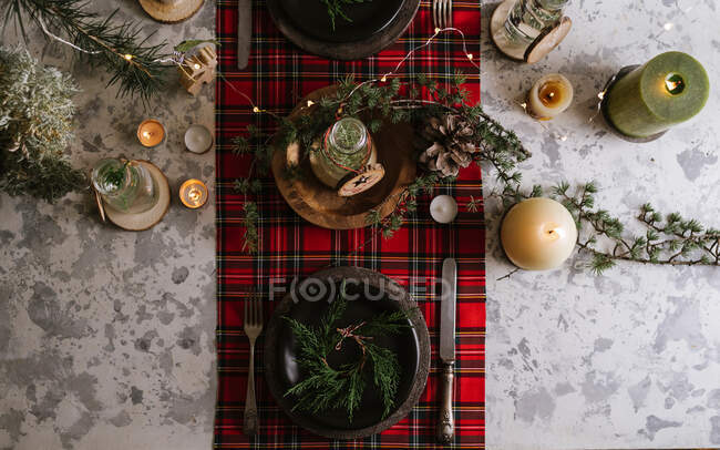Top view of Christmas table setting with wreath on the plate, decorative wooden ornaments and red checkered tablecloth with yellow lights on the background — Stock Photo