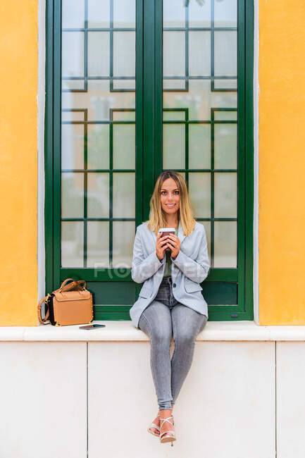 Ganzkörper zufriedene Frau im trendigen Outfit mit Pappbecher Kaffee blickt in die Kamera, während sie mit Handtasche in der Nähe des Gebäudes sitzt — Stockfoto