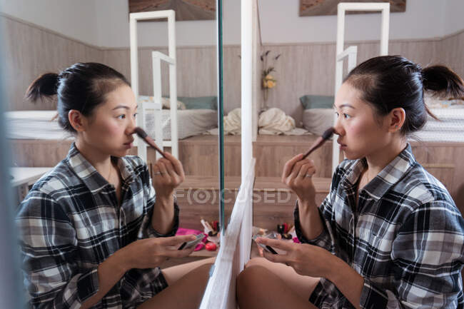 Вид збоку молодої етнічної жінки, що сидить біля дзеркала, прикладаючи порошок у світлій спальні з ліжком і столом з комп'ютером — стокове фото