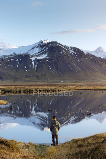 Обратный вид на неузнаваемого туриста-мужчину в повседневной одежде, любующегося дикой природой, стоя возле мирного озера, отражающего снежные горы Исландии — стоковое фото