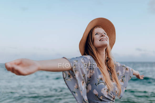 Усміхнена молода жінка в повсякденному одязі, що стоїть на піщаному пляжі біля моря, коли влітку з відкритими руками — стокове фото