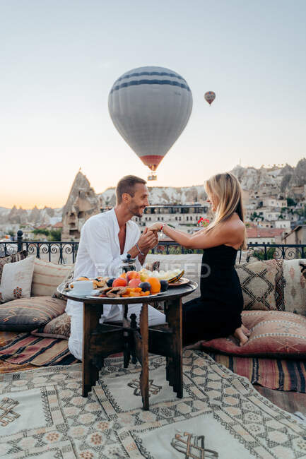Homme embrassant la main de la petite amie tout en étant assis sur le toit en ville avec des montgolfières volant dans le ciel du soir — Photo de stock